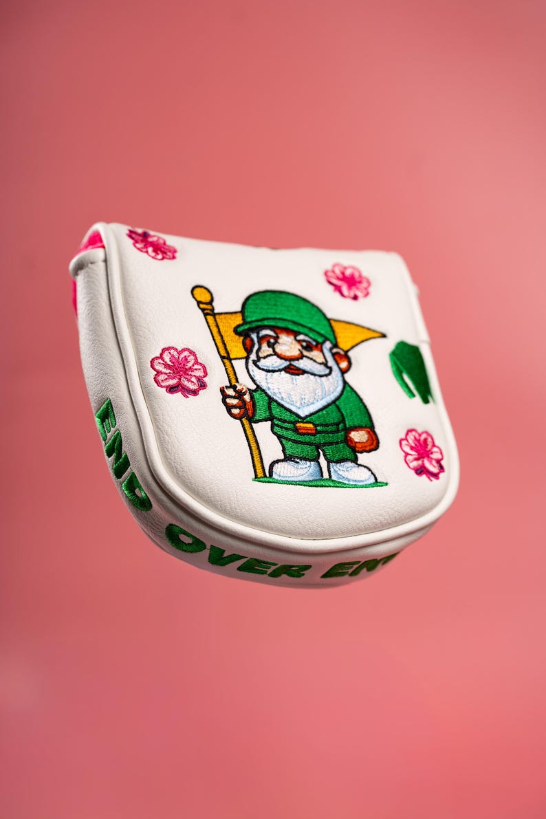 Puede incluir: Una funda blanca para putter de golf con un gnomo bordado vestido de verde que sostiene una bandera amarilla. El dise&ntilde;o incluye detalles de flores rosas y el texto bordado en verde END OVER END a lo largo del borde lateral de la funda.