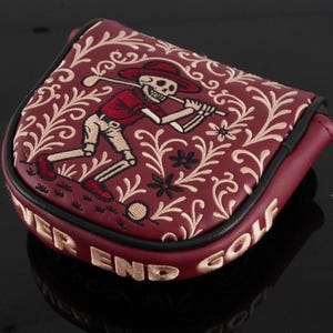Puede incluir: Funda de putter de golf granate con un diseño de golfista esqueleto. La funda presenta un esqueleto bordado con sombrero y camisa rojos, balanceando un palo de golf. Las palabras "NEVER END GOLF" están bordadas en dorado.