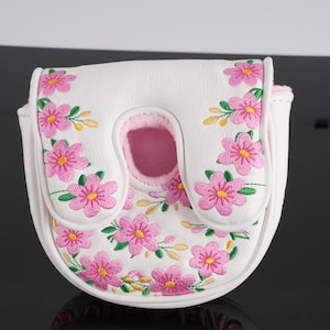 May include: A white mallet-style golf putter headcover featuring an embroidered pattern of pink flowers with yellow centers and green leaves. The cover has a U-shaped cutout at the top and a light pink interior lining visible through the opening.