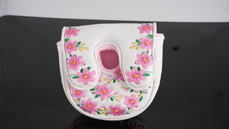May include: A white mallet-style golf putter headcover featuring an embroidered pattern of pink flowers with yellow centers and green leaves. The cover has a U-shaped cutout at the top and a light pink interior lining visible through the opening.