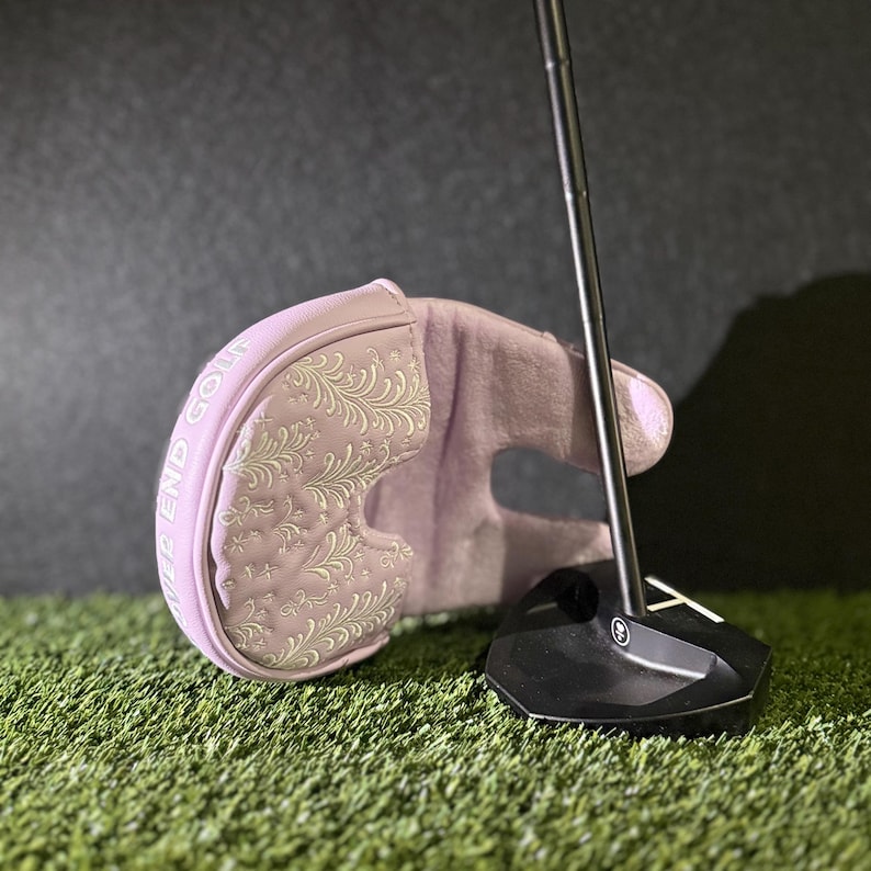 May include: A light purple golf putter head cover with an embroidered floral design and the words "River End Golf". The cover is resting on a bed of green grass next to a black golf club.
