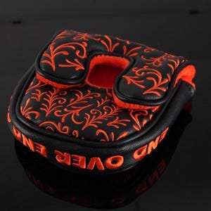 May include: A black mallet putter headcover featuring an orange embroidered floral vine pattern on the top surface. The side of the headcover displays the orange embroidered text END OVER END, and the interior is lined with bright orange fabric.
