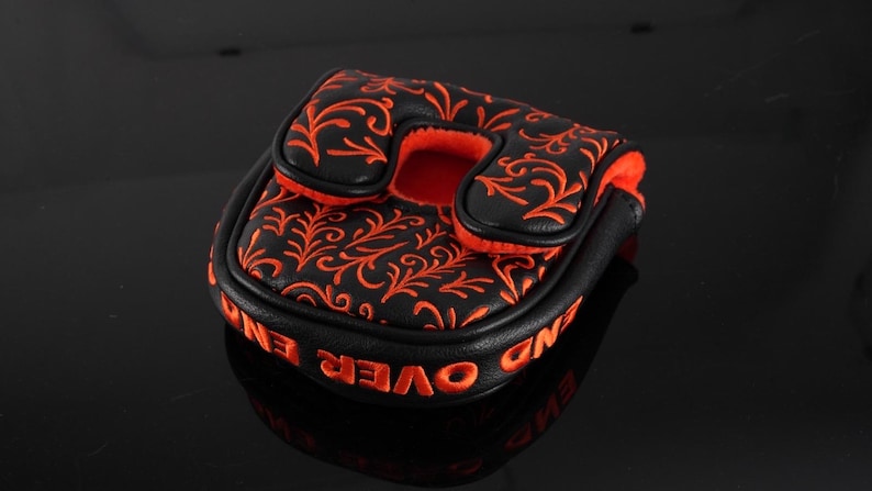 May include: A black mallet putter headcover featuring an orange embroidered floral vine pattern on the top surface. The side of the headcover displays the orange embroidered text END OVER END, and the interior is lined with bright orange fabric.