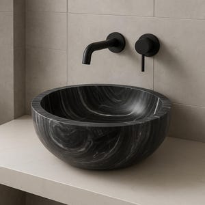 Peut inclure: Un lavabo en marbre noir avec un motif tourbillonnant blanc repose sur un comptoir de couleur claire. Au-dessus, un robinet noir mat et une poignée assortie sont fixés au mur. L'évier est rond et a une finition lisse et polie.