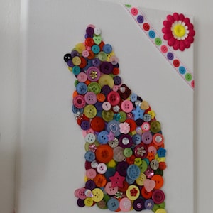 May include: A colorful cat silhouette created with buttons on a white canvas. The cat is outlined with buttons in various colors, including red, blue, green, yellow, and purple. The canvas is decorated with pink and white ribbon.