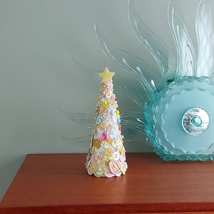 May include: A small, cone-shaped Christmas tree made of colourful buttons. The tree is decorated with a yellow star on top and is sitting on a wooden surface.