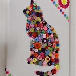 May include: A white canvas with a colorful button cat design. The cat is made of various sizes and colors of buttons, including red, blue, green, yellow, and pink. The canvas is decorated with a pink flower and a colorful ribbon.