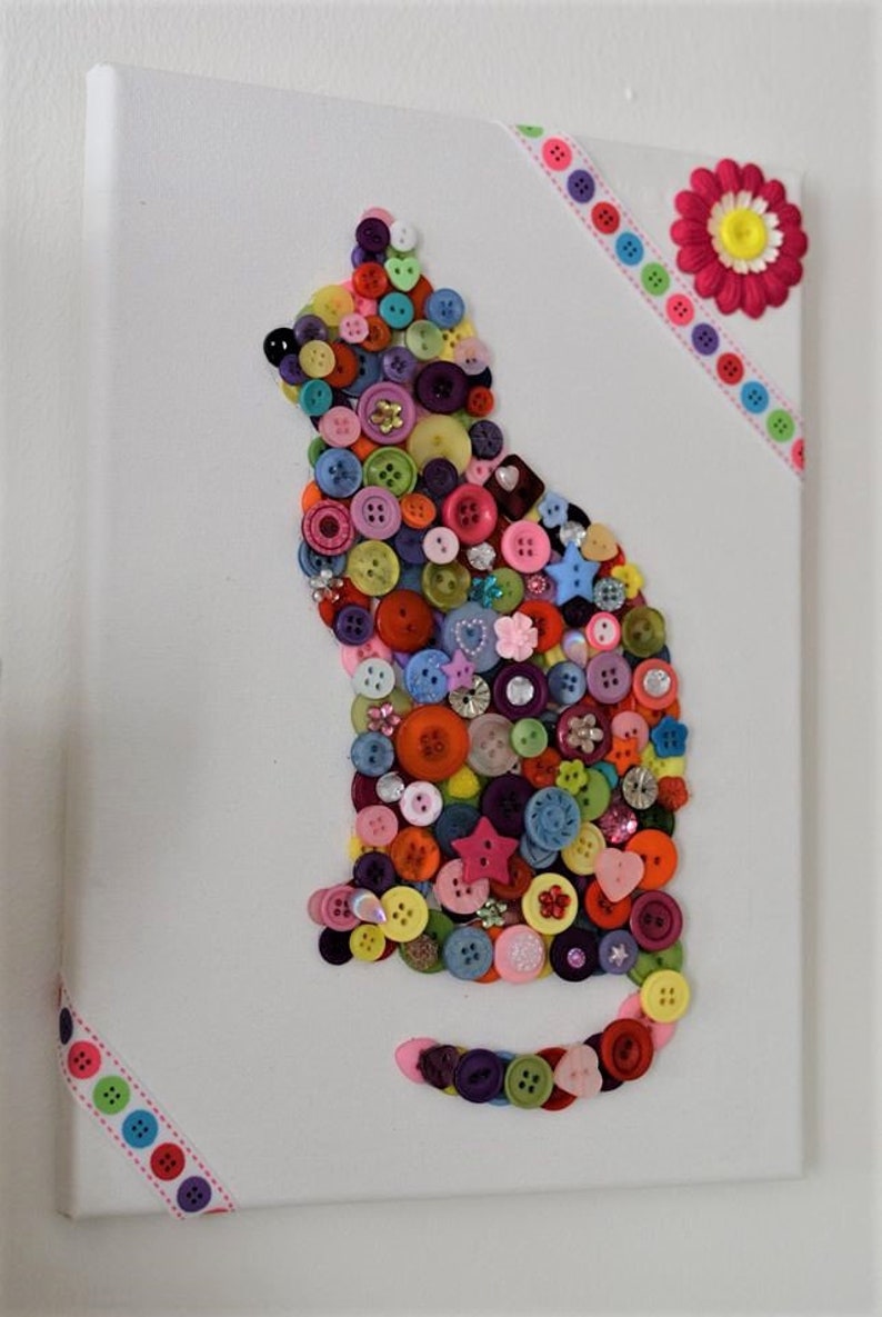 May include: A white canvas with a colorful button cat design. The cat is made of various sizes and colors of buttons, including red, blue, green, yellow, and pink. The canvas is decorated with a pink flower and a colorful ribbon.