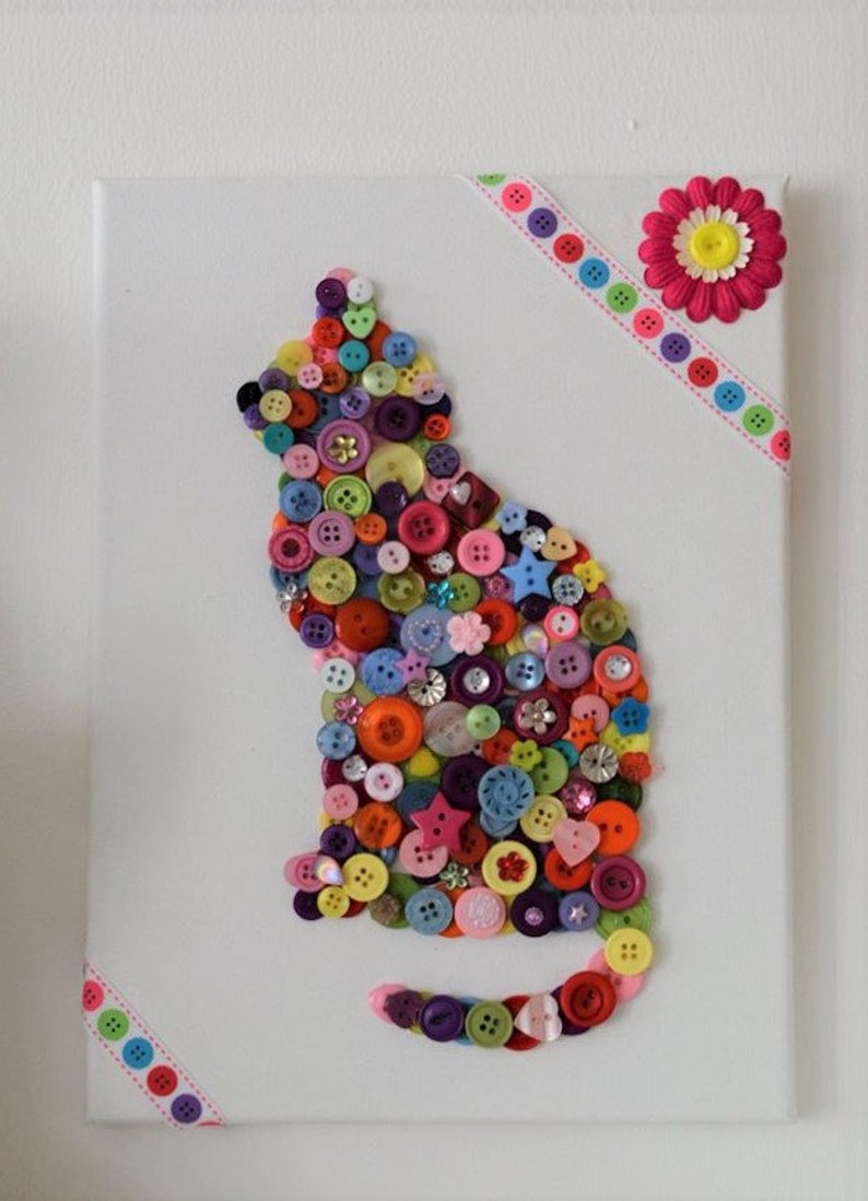 May include: A white canvas with a colorful button cat design. The cat is made of various colored buttons, including red, blue, green, yellow, and purple. The canvas is decorated with pink and white ribbon.