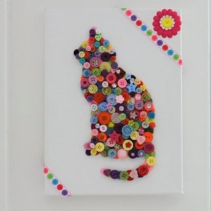 May include: A colorful cat silhouette made from buttons on a white canvas. The cat is surrounded by a pink and white ribbon with a pink flower in the top right corner.