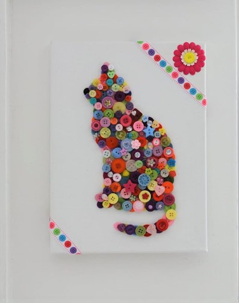 May include: A colorful cat silhouette made from buttons on a white canvas. The cat is surrounded by a pink and white ribbon with a pink flower in the top right corner.