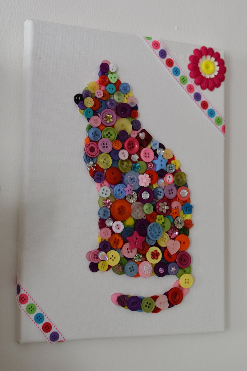 May include: A colorful cat silhouette created with buttons on a white canvas. The cat is outlined with buttons in various colors, including red, blue, green, yellow, and purple. The canvas is decorated with pink and white ribbon.