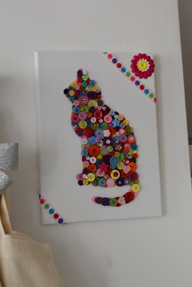 May include: A colorful cat silhouette made from buttons on a white canvas with a pink and white flower and colorful ribbon accents.