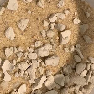 May include: Close-up of a container filled with a mixture of light tan sand and irregularly shaped, off-white, rock-like pieces. The sand appears granular, and the rocks vary in size and texture.