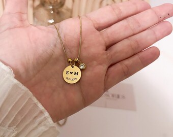 以下が含まれることがあります： イニシャルと日付が刻まれた丸いペンダントと小さなクリスタルチャームが付いたゴールドカラーのネックレス。画像には「PERFECT GIFT、PERSONALIZED INITIAL + DATE、CUSTOM MADE、SHIPS FAST」というテキストが含まれています。背景は柔らかいです。