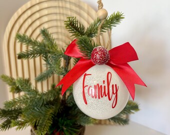 以下が含まれることがあります： 織りバスケットに入った小さなクリスマスツリー。赤い文字で「Family」と書かれた白いオーナメントで飾られています。オーナメントは赤いリボンと装飾的なベリーでアクセントがつけられています。背景には木のアーチがあります。