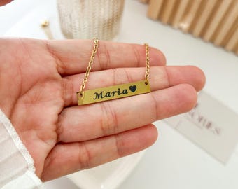 以下が含まれることがあります： 長方形のペンダントが付いたゴールドカラーのネックレス。ペンダントには「Maria」という名前が黒い筆記体で刻まれ、小さな黒いハートのシンボルが続きます。ネックレスは手の上に表示されています。