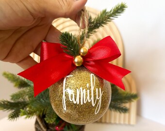 以下が含まれることがあります： 白い筆記体で「family」の文字が入った、金色のグリッタークリスマスオーナメント。赤いサテンのリボン、金のビーズ、吊り下げ用の紐ループで飾られています。ホリデーデコレーションの一部です。