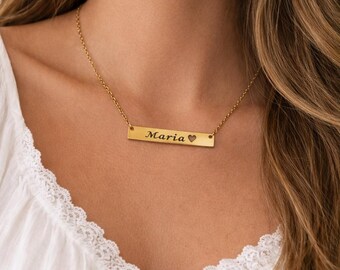 以下が含まれることがあります： 黒で「Maria」という名前が刻まれた金のバーネックレスと小さなハートのシンボル。ネックレスは繊細なチェーンから吊り下げられています。白いトップスを着ています。