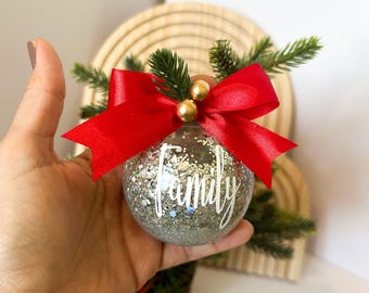 以下が含まれることがあります： 銀色のグリッターが詰められた透明なクリスマスオーナメントで、白いスクリプトで「Family」という文字が書かれています。赤いサテンのリボン、人工の常緑樹の小枝、金色のビーズのアクセントで飾られています。お祝いの休日の装飾。