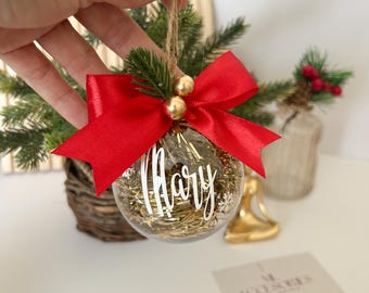 以下が含まれることがあります： 金色のきらびやかな飾りで満たされた透明なクリスマスオーナメント。白い筆記体で「Mary」という名前が書かれています。赤いサテンのリボン、金のビーズ、麻ひものハンガーが付いています。小さな常緑樹の小枝と雪の結晶も見えます。