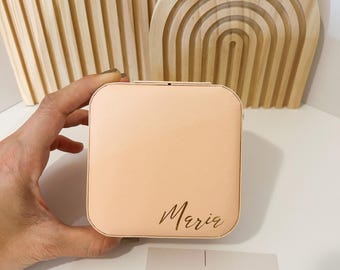 以下が含まれることがあります： 前面に「Maria」と刻印された、桃色の正方形のジュエリーボックス。 手に持たれており、白い表面に置かれています。 「MIL ACCESORIES」というテキストのビジネスカードも表示されています。