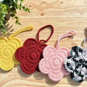 May include: Four crocheted flower-shaped ornaments with hanging loops. The flowers are solid yellow, deep red, light pink, and a variegated black, white, and gray pattern. Each piece features a circular center with five rounded petals.