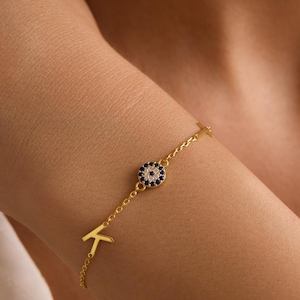 May include: A fine gold bracelet with a blue and white evil eye pendant and the letter "K". The bracelet is displayed on a wrist. The charm is encircled by tiny blue gemstones.