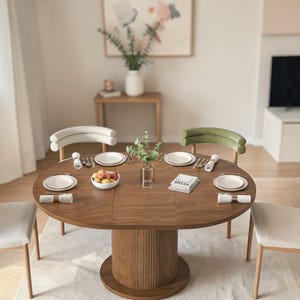 May include: A round wooden dining table with a pedestal base, set for a meal. The table is surrounded by chairs with neutral-colored upholstery. The table is set with plates, silverware, napkins, and a bowl of fruit. A vase of greenery and a small book are also on the table.
