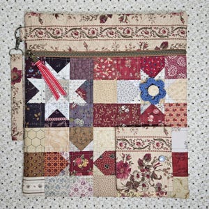 May include: A patchwork zippered pouch with a wrist strap. The design features squares of various floral and geometric patterns in red, cream, and tan. A blue crocheted flower and a red ribbon accent the front. The pouch has a metal zipper and a cream-colored floral border.
