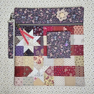 May include: A quilted clutch with a patchwork design featuring floral and star patterns in shades of purple, red, and cream. It has a zipper closure, a wrist strap, and a small pocket. A crocheted flower embellishes the top.