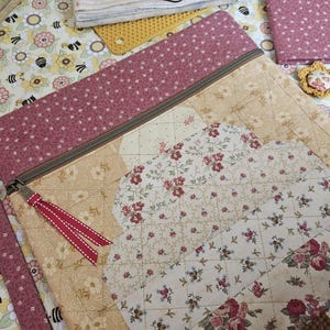 May include: A quilted zipper pouch featuring a patchwork design of floral fabrics in shades of dusty rose, cream, and tan. The pouch has a metal zipper with a red and white striped ribbon pull and a matching fabric wrist strap attached to the side.