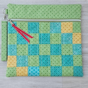 May include: A quilted pouch with a patchwork design in shades of green, teal, and yellow. The pouch has a zipper closure with a red ribbon pull and a wrist strap. The fabric features a repeating pattern of small crosses.