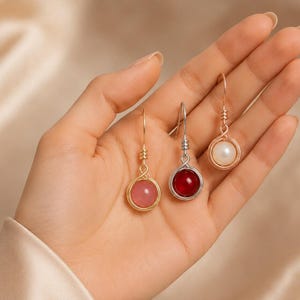 May include: Three handmade earrings with wire-wrapped gemstone and pearl pendants. The earrings feature a pink gemstone, a red gemstone, and a white pearl, each suspended from a delicate wire hook. The metal is gold and silver.