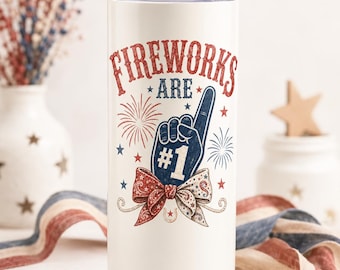 May include: A white cylindrical tumbler with a metal straw and a lid. The design features the red text FIREWORKS ARE, a blue foam finger with the number 1, red and blue firework bursts, small stars, and a patterned bow in red, white, and blue.