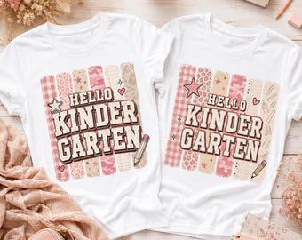 May include: Two white t-shirts featuring a graphic print with the text "HELLO KINDERGARTEN" in a bold font. The design includes vertical stripes with pink patterns, stars, hearts, and a pencil illustration. The shirts have short sleeves and crew necks.