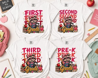 May include: Four white t-shirts featuring a graphic of a raccoon wearing glasses and a bow, holding a pencil and book. The designs include the text First Grade, Second Grade, Third Grade, and Pre-K, along with stacks of books and crayons.