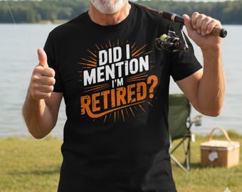 May include: Black t-shirt with the text "DID I MENTION I'M RETIRED?" in white and orange. The shirt is worn by a person giving a thumbs up and holding a fishing rod. The background shows a lake and green grass.