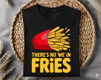 May include: Black t-shirt featuring a graphic of a red carton overflowing with golden french fries. The text "THERE'S NO 'WE' IN FRIES" is in yellow. The shirt is folded on a woven basket.