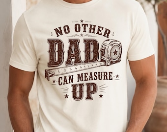 May include: A cream-colored short-sleeve t-shirt featuring a brown graphic print. The design includes the text "NO OTHER DAD CAN MEASURE UP" surrounding an illustration of a tape measure. The graphic is centered on the chest of the garment.