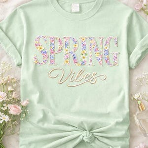 Peut inclure: T-shirt vert clair avec l'inscription "SPRING Vibes" en motif floral. Le t-shirt est noué à la taille. Des fleurs et des perles sont disposées autour du t-shirt.