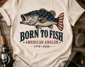 May include: A cream-colored t-shirt featuring a graphic of a fish with a blue and white star-patterned body and red and white striped tail. Below the fish, the text reads BORN TO FISH, AMERICAN ANGLER, and the years 1776 and 2026.