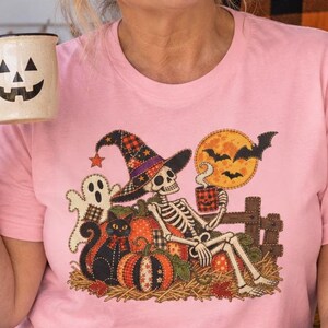 Op de afbeelding: Een lichtroze T-shirt met een grafische print van een skelet met een geruite heksenhoed, zittend tussen pompoenen en een zwarte kat. Een geest staat in de buurt en op de achtergrond is een volle maan met vleermuizen te zien.