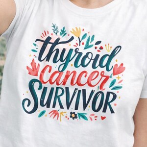 May include: White t-shirt with the words "thyroid cancer survivor" in a colorful floral design. The shirt is being held up by a person smiling at the camera. A purple, pink, and blue ribbon is in the background.