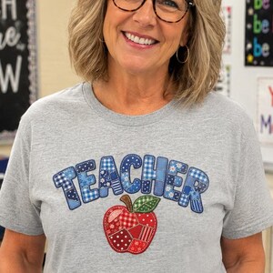 Op de afbeelding: Een grijs gemêleerd T-shirt met korte mouwen en het woord TEACHER in blauw geruite en bedrukte letters boven een rode appelafbeelding gemaakt van diverse patchwork-stofontwerpen. Het shirt wordt gedragen met een blauwe spijkerbroek en een zilverkleurige armband.