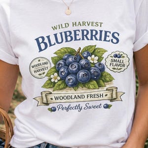 May include: A white t-shirt featuring a graphic of blueberries with green leaves and text that reads Wild Harvest Bluberries, Woodland Harvest, Small Flavor, Woodland Fresh, and Perfectly Sweet, along with small illustrations of individual blueberries.