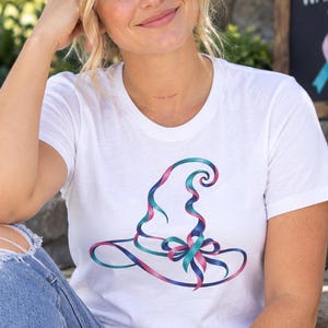 May include: A white short-sleeve t-shirt featuring a graphic of a witch hat formed by a swirling teal and pink ribbon. The ribbon design includes a central bow detail. The shirt is made of a plain white fabric with a crew neckline and short sleeves.