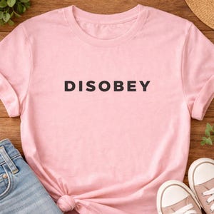 May include: A light pink t-shirt with the word "DISOBEY" printed in black block letters. The shirt is made of soft material and is styled with a knot at the bottom. Blue jeans and pink sneakers are visible.