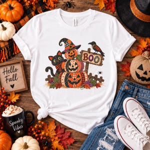 Può includere: Una t-shirt bianca a maniche corte con una stampa grafica di tre zucche impilate, un gatto nero e un corvo appollaiato su un cartello di legno con la scritta BOO. Il design presenta un motivo patchwork con colori vivaci.
