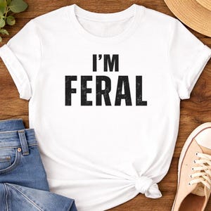 May include: White t-shirt with the text "I'M FERAL" in bold, black letters. The shirt is knotted at the waist. Blue jeans, a straw hat, and beige sneakers are partially visible.